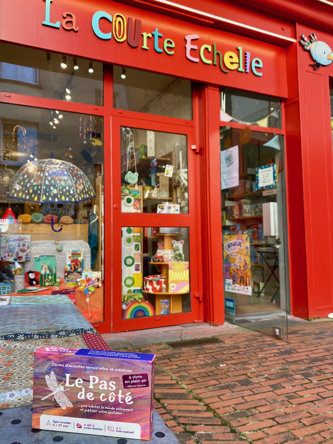 Vitrine de la librairie jeunesse et magasin de jouets La Courte Échelle à Rennes et jeu de cartes Le Pas de côté au premier plan